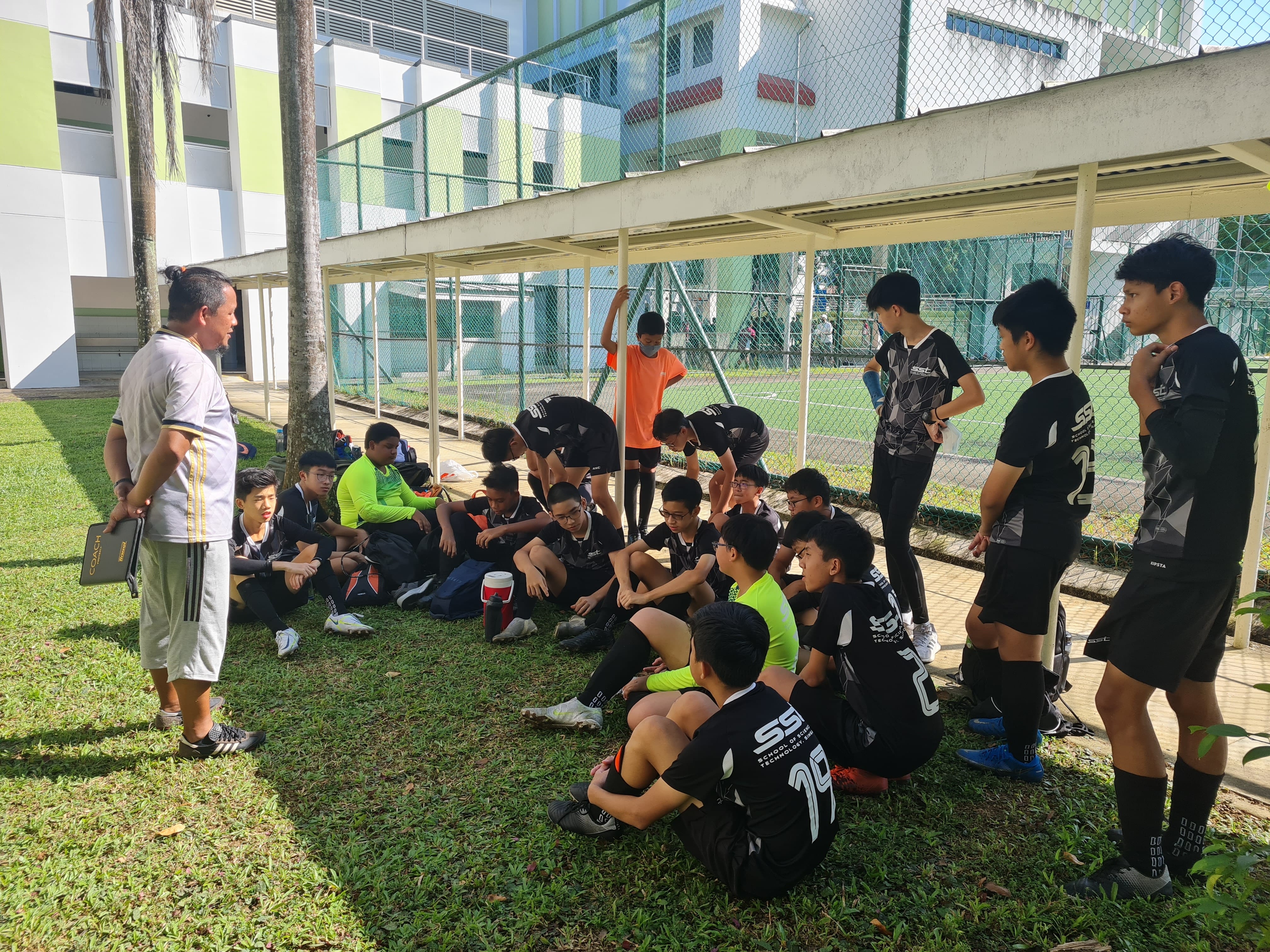 Coach talks to seated soccer players in black uniforms resting beside a fenced field and school building